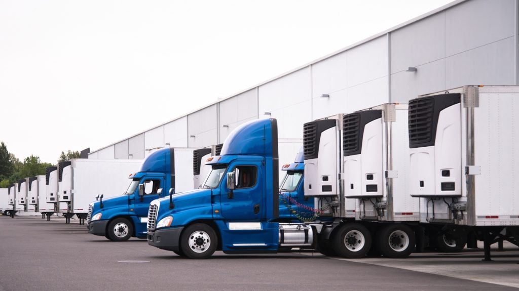 A day cab big rigs semi trucks with a reefer trailers stand near the gate of the warehouse next to other reefer trailers that are loaded and unloaded to deliver perishable and frozen food to consumers